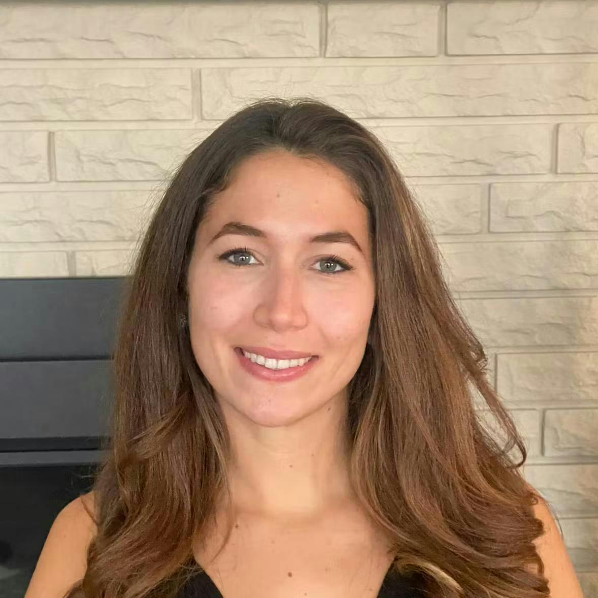 Woman smiling against a beige brick wall background. She has long, wavy, brown hair and is wearing a black top.