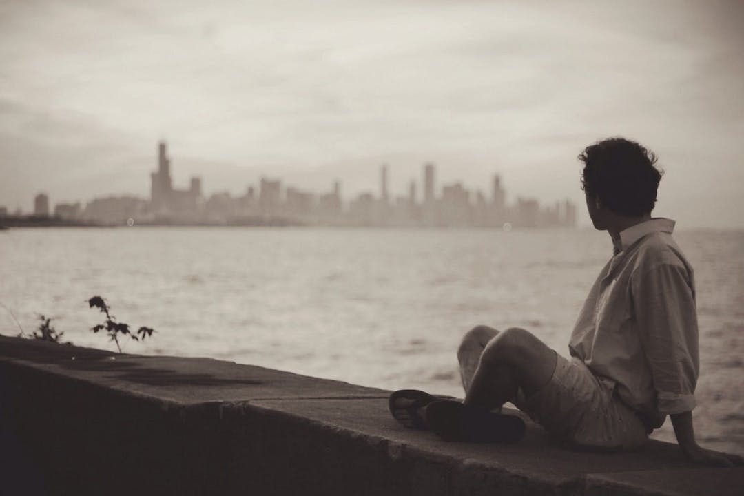 A person sits on a ledge looking over a large body of water, with a city skyline in the distant background under a cloudy sky.