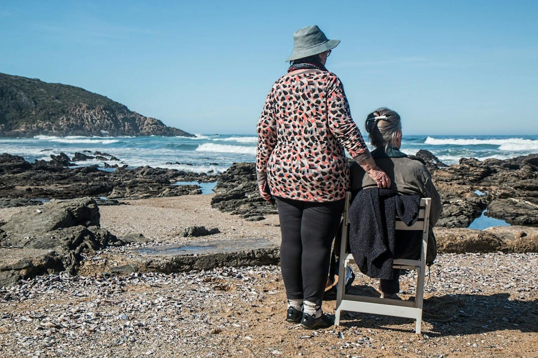 Two women, one standing with a hand on the other's chair, face a rocky beach with waves and a distant hill under a clear blue sky.