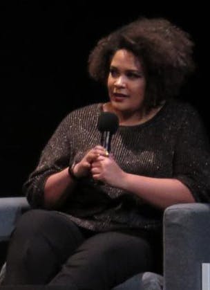 A person holding a microphone speaks while seated on a stage, wearing a dark, sparkly top and black pants. The background is black, and there is another seat next to them.
