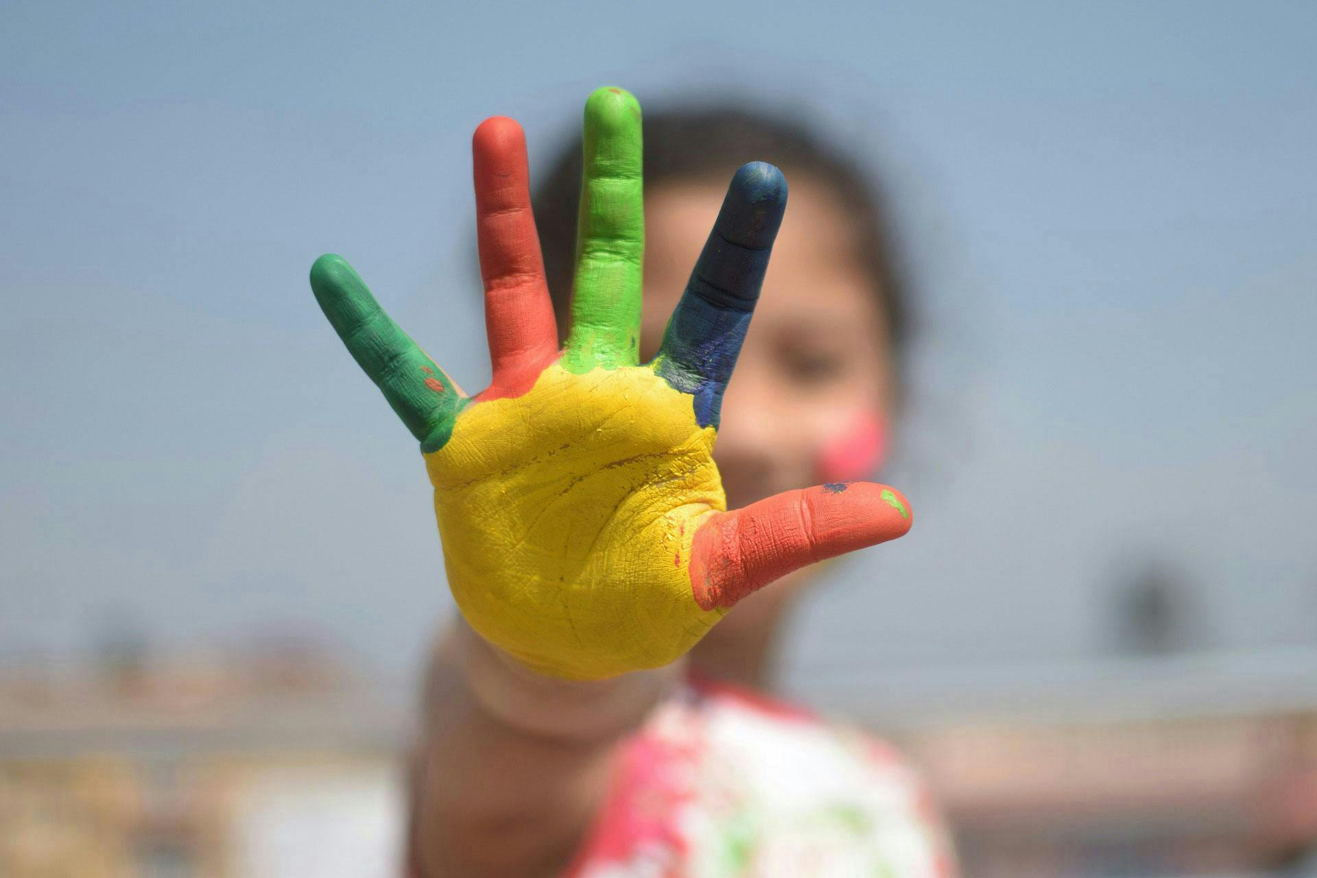 A hand painted in rainbow colors (red, green, yellow, blue) is outstretched towards the camera. A child blurred in the background forms the context on a sunny, outdoor day.