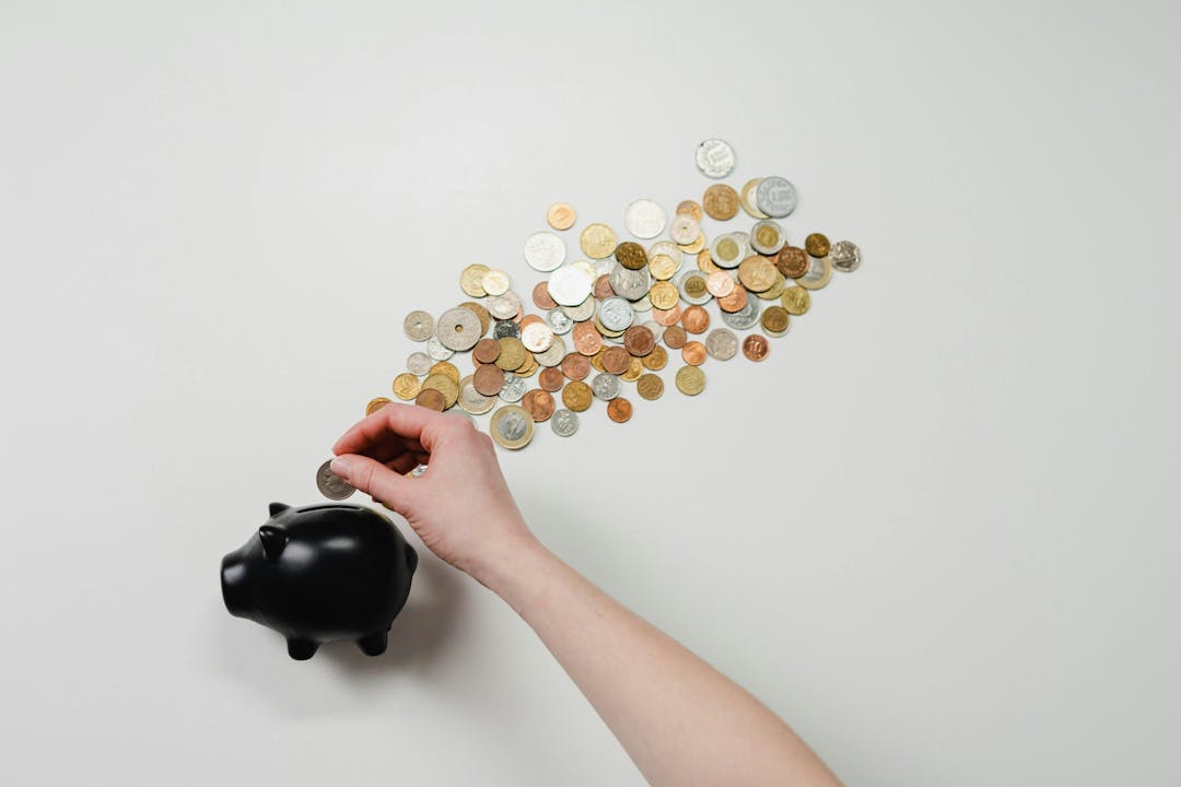 A hand inserts a coin into a black piggy bank, surrounded by a scattered pile of various coins on a white surface.