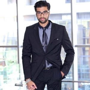 A man in a black suit and tie stands confidently with one hand in his trouser pocket, in front of a large window overlooking a cityscape.