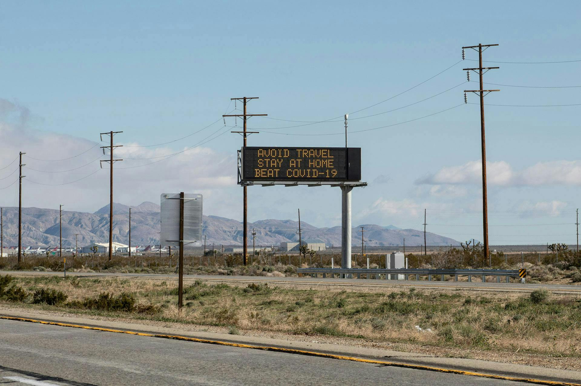 Digital highway sign displays "AVOID TRAVEL STAY AT HOME BEAT COVID-19" in a desert landscape, with a distant mountain range and scattered power lines.