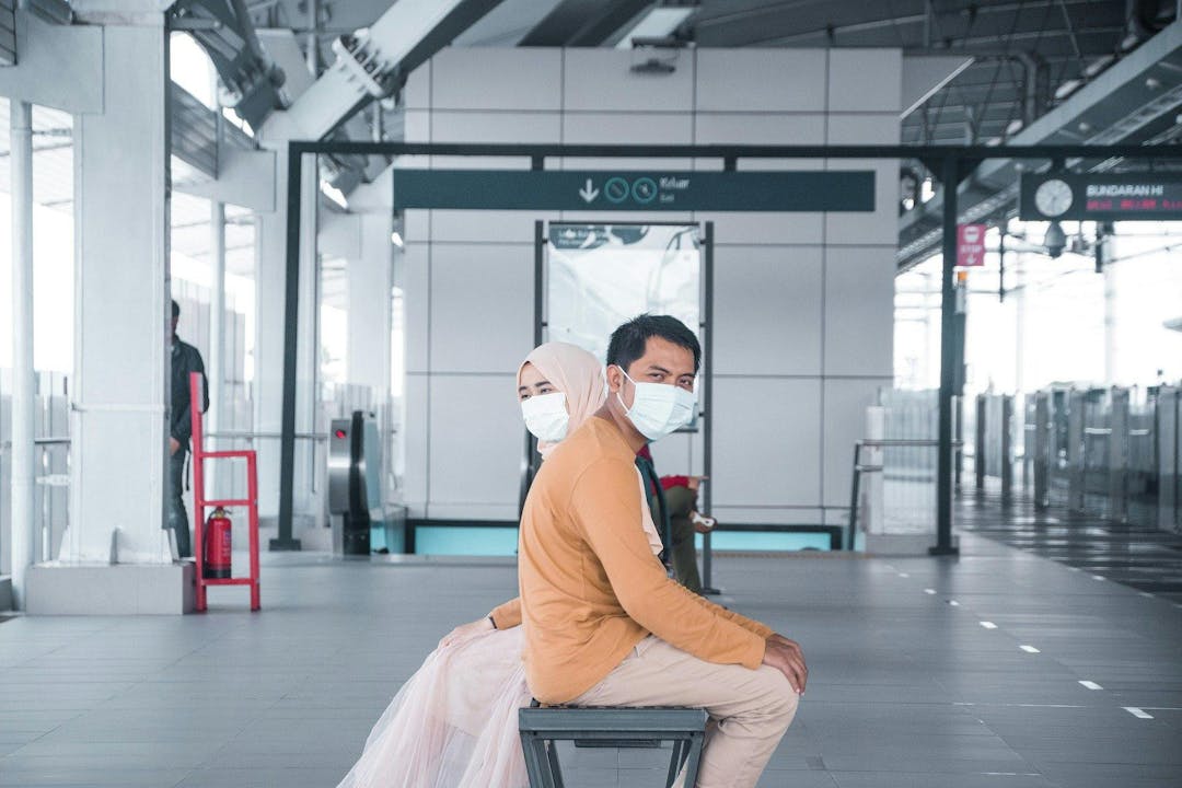 A masked couple sits back-to-back on a bench in a modern, open train station. Signs indicate "Keluar Exit" and "Blok G," with other passengers and structural beams in the background.