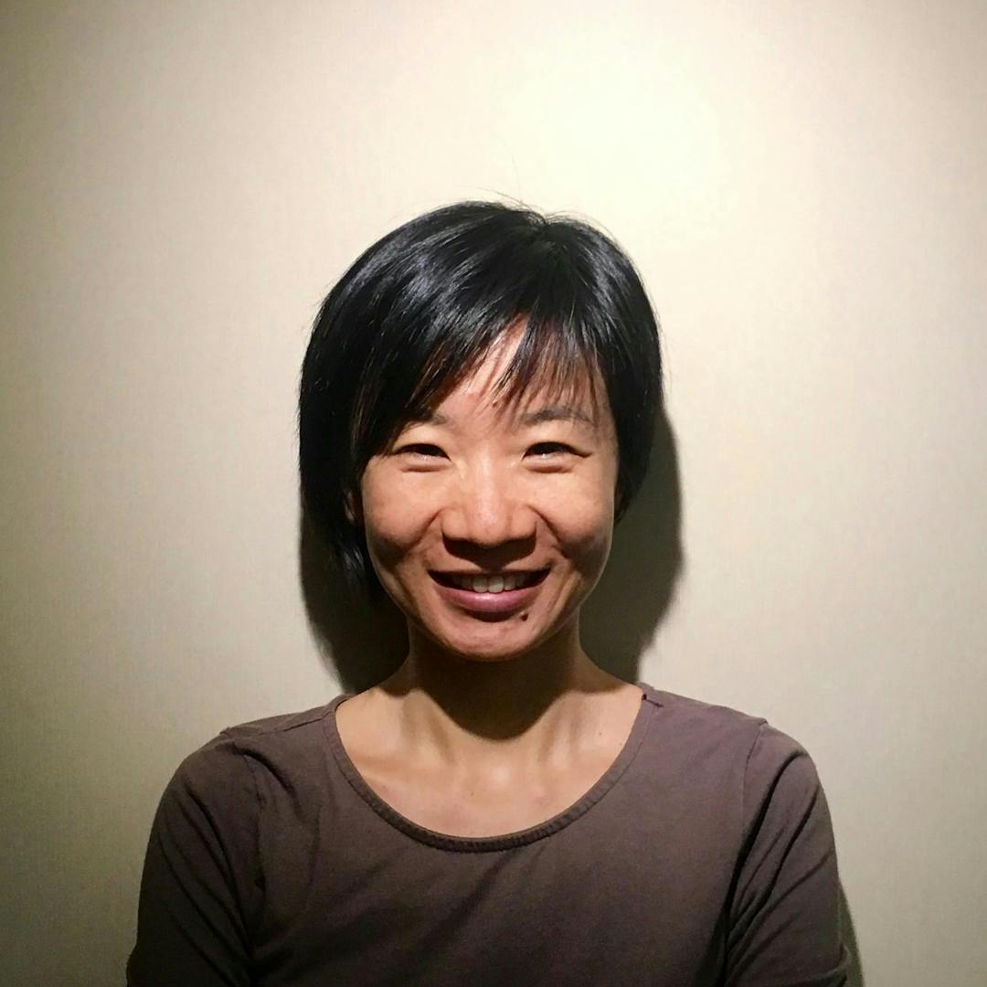 A person, smiling gently, stands against a plain, beige background, wearing a brownish-gray top, with short, black hair casting a slight shadow on the wall.