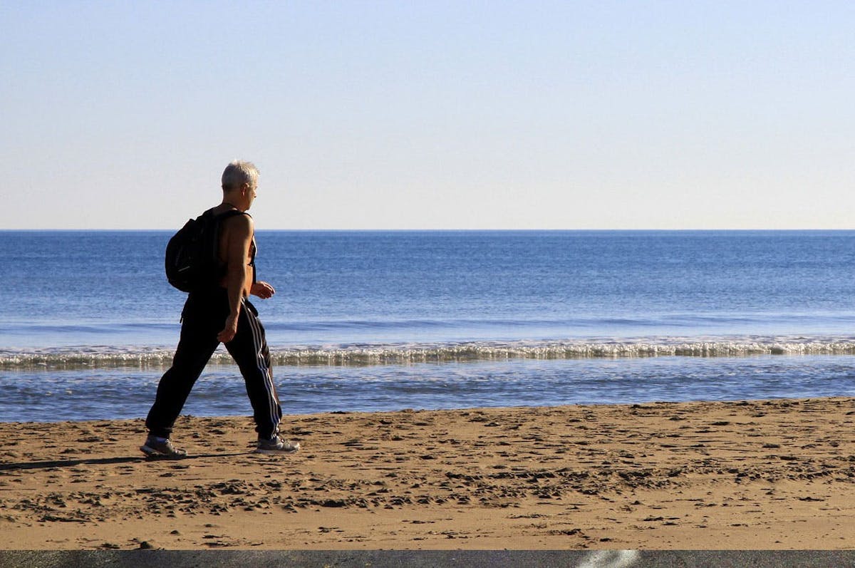 Person walking on sandy beach beside calm ocean, wearing black pants, tank top, and carrying a backpack, under a clear, blue sky.