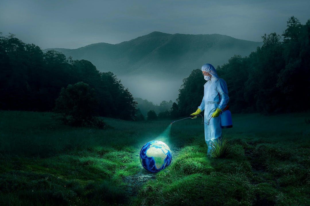A person in protective gear sprays a globe lying on the grass in a misty, mountainous landscape at twilight.