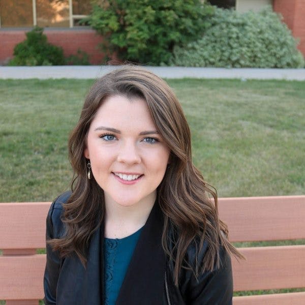 A young woman with long, wavy hair sits on a bench, smiling. She wears a blue top and a dark jacket. The grassy courtyard and a brick building with windows and greenery are in the background.