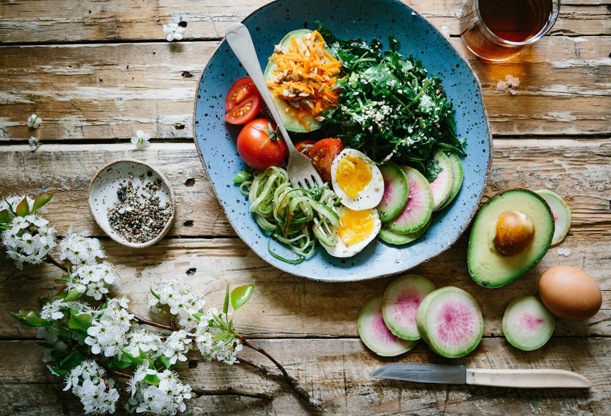 A blue plate holds a colorful salad with eggs, avocado, and vegetables; placed on a rustic wooden table with sliced radishes, an avocado half, a boiled egg, and a small bowl of pepper.