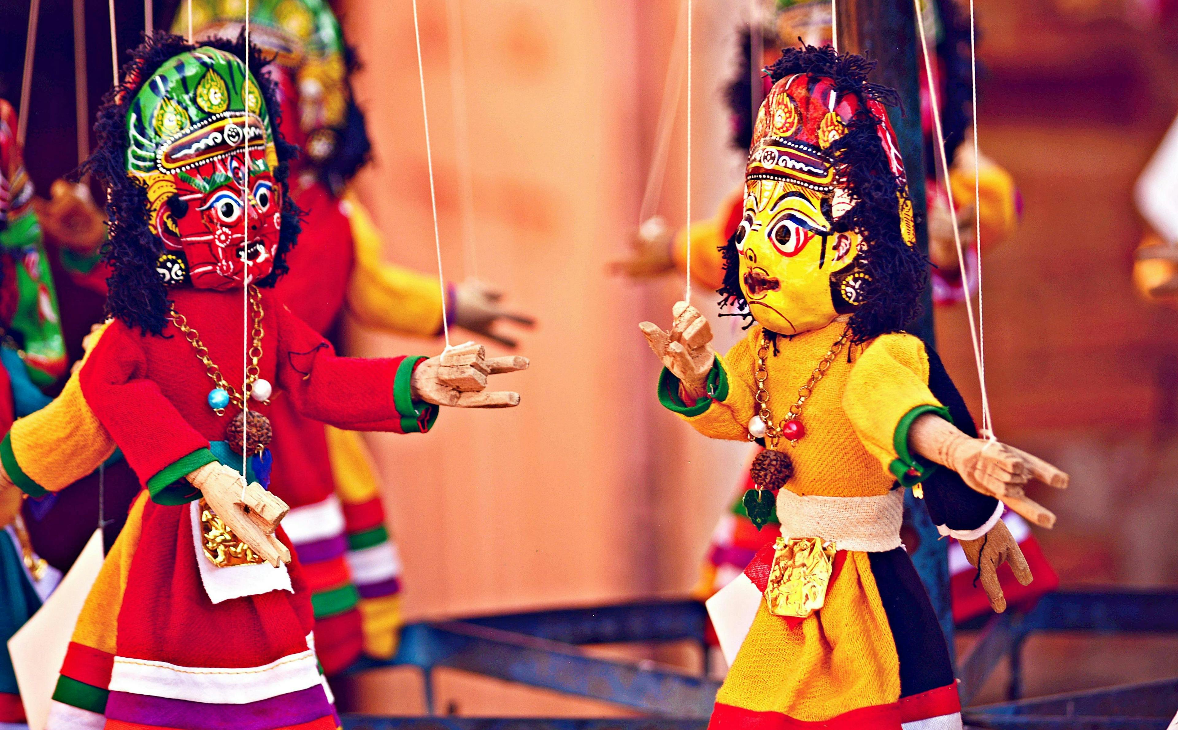 Colorful traditional puppets with intricate face paint and elaborate costumes, suspended by strings, appear to be engaging in a dynamic performance. The background is softly blurred, highlighting the puppets.