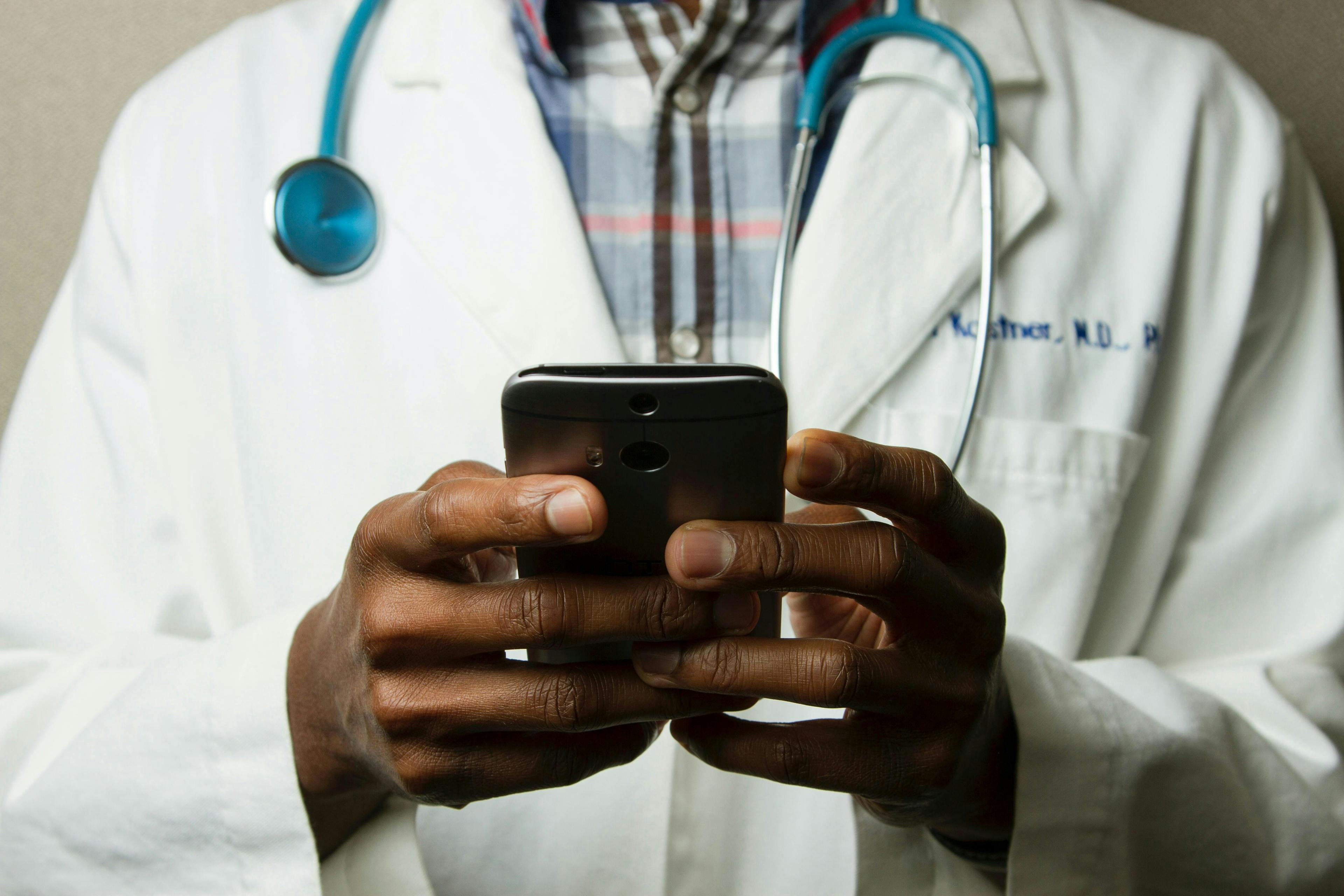 A person in a white lab coat with a stethoscope uses a smartphone; the background is a plain, neutral color.