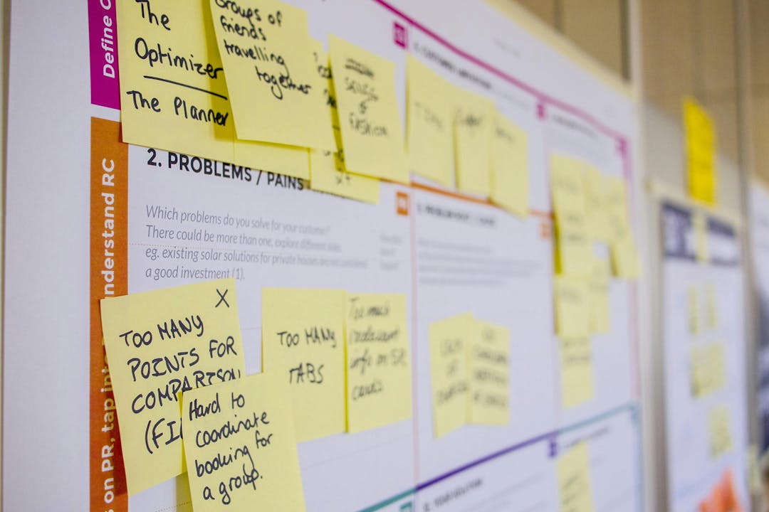 Sticky notes containing handwritten text are attached to a whiteboard in a brainstorming session, highlighting various problems.

Visible text: 
"Too many points for comparison (FI)",
"Hard to coordinate booking for a group",
"Groups of friends travelling together",
"Too many tabs".