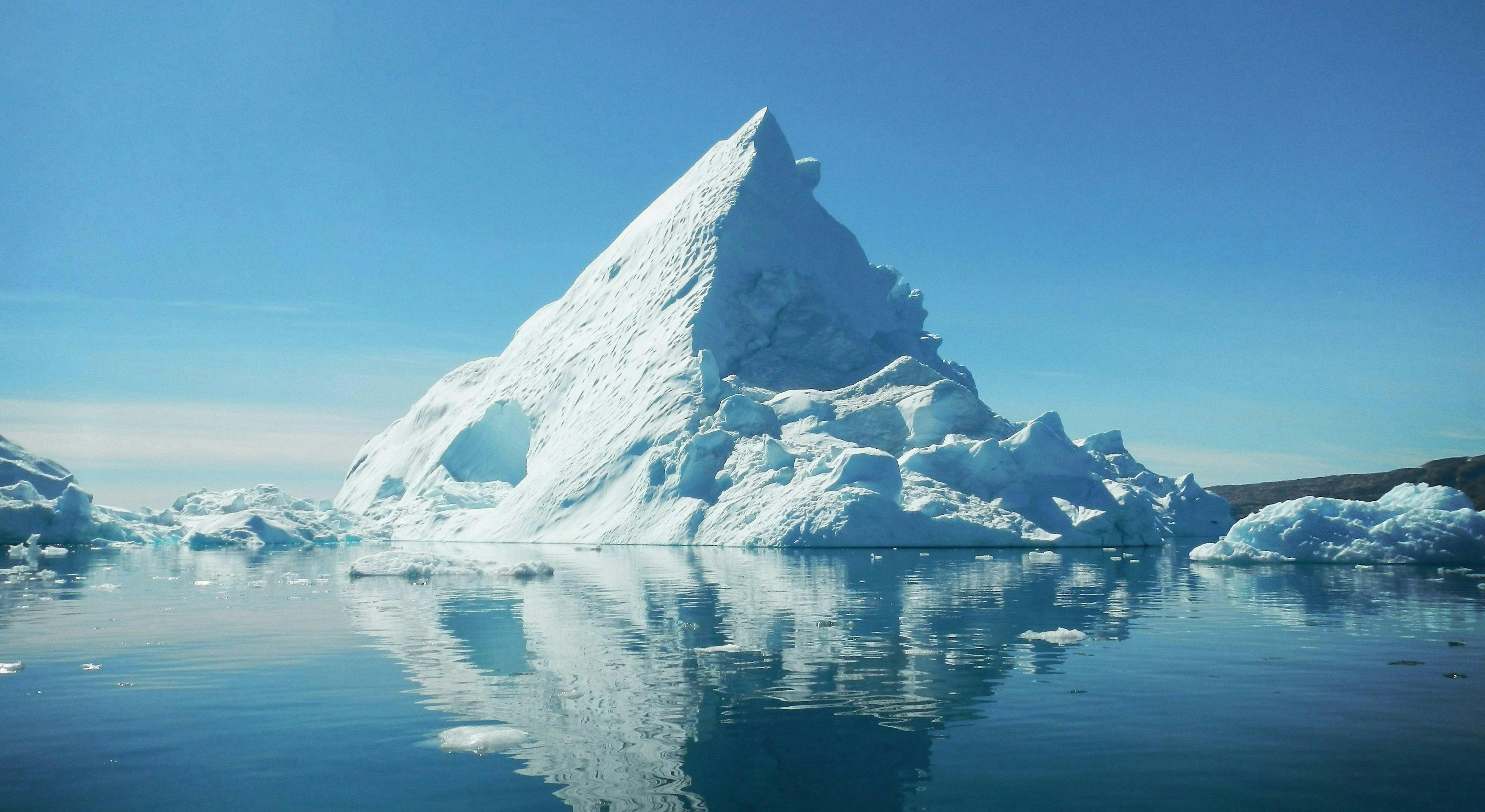 Large iceberg reflecting in calm, clear arctic waters under a bright blue sky, surrounded by smaller ice formations, suggesting a cold, remote environment.