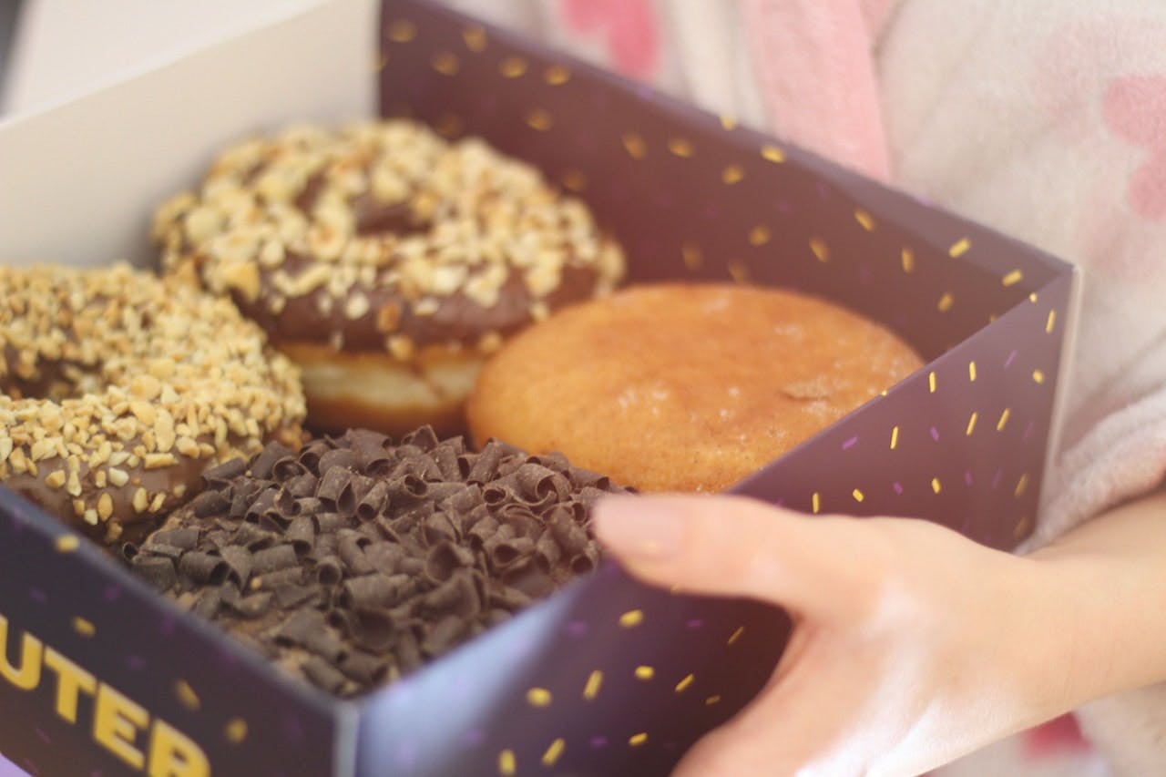 A hand holds a box containing four donuts with various toppings; chocolate sprinkles, crushed nuts, plain, in a cozy setting.