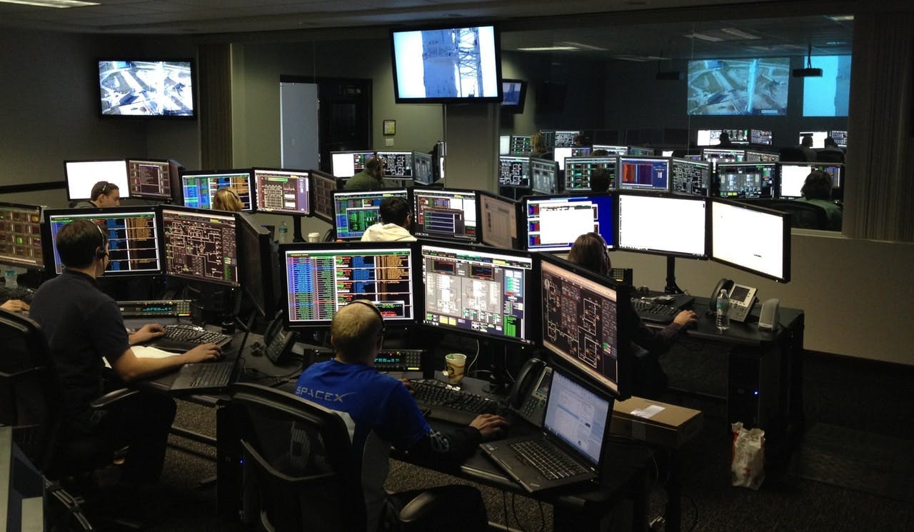 A group of people operates numerous computer monitors at workstations in a control room with wall-mounted screens displaying data and images.