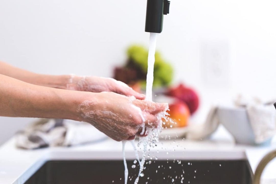 Hands cleansing under running water; the kitchen sink area surrounds with out-of-focus fruits and cloths in the background.