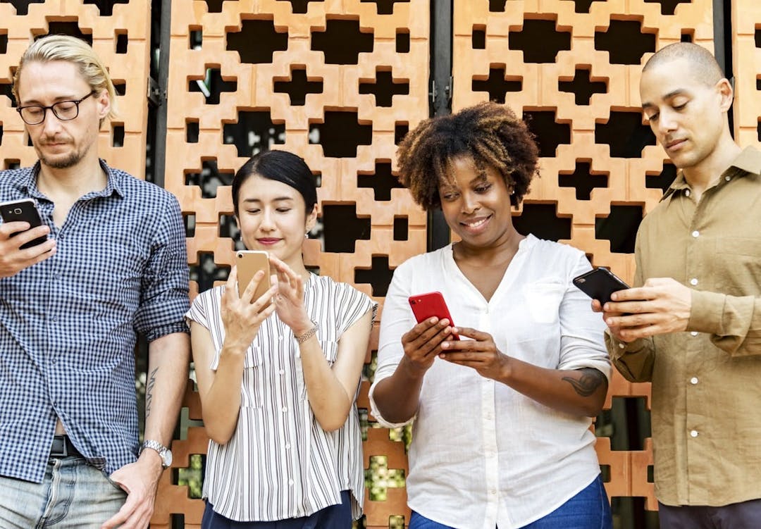 Four people stand against a patterned brick wall, all looking down and engaging with their smartphones. The context appears to be an outdoor setting with a decorative background.