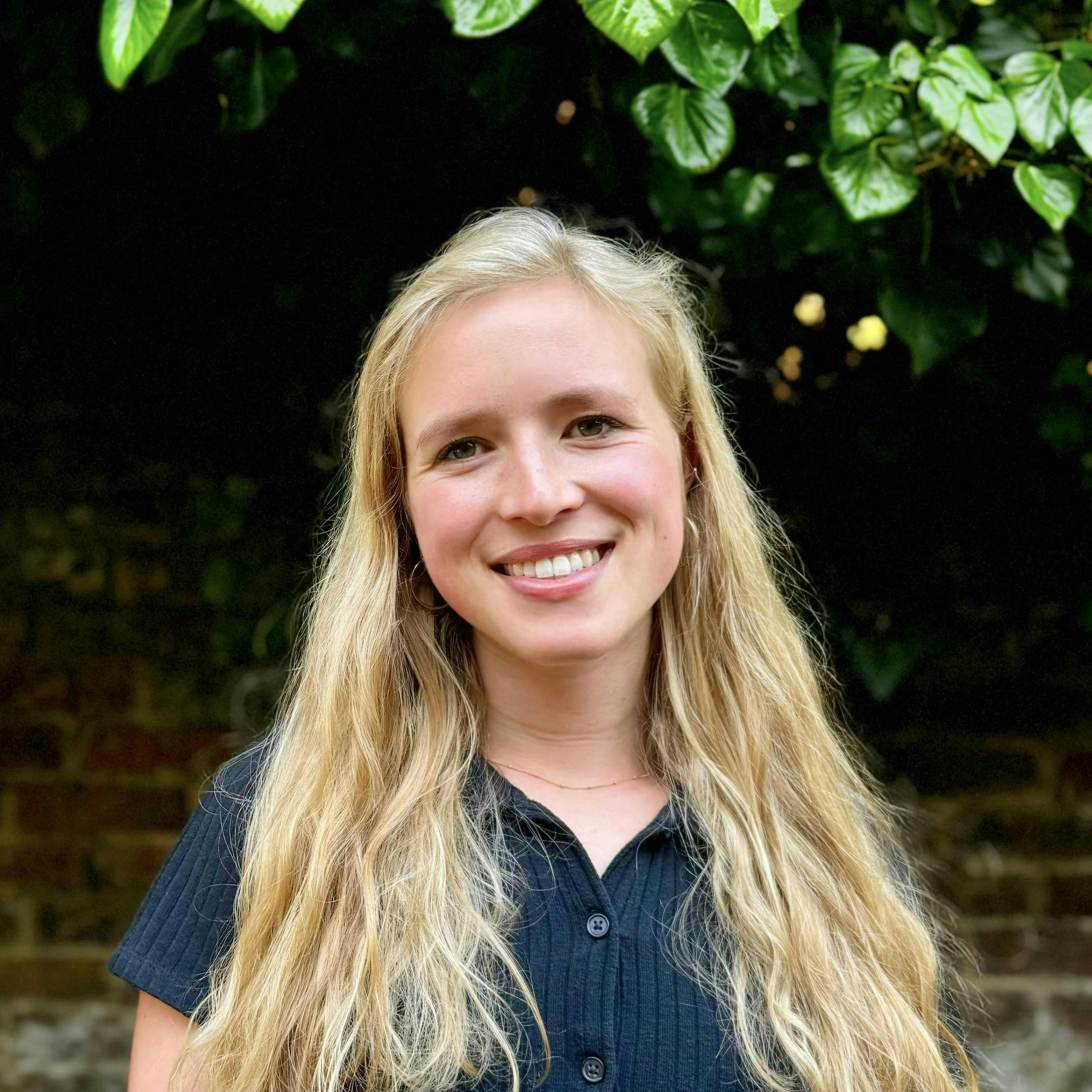 A smiling woman with long blonde hair is standing, wearing a dark button-up shirt, set against a backdrop of green foliage and a brick wall.