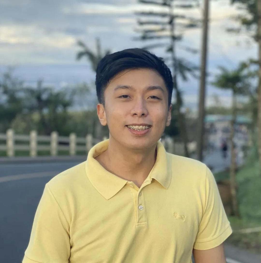 The image shows a  man with short, dark hair, wearing a yellow collared shirt, smiling at the camera. He is standing outdoors near a road with a scenic background featuring trees, a fence, and a slightly cloudy sky.