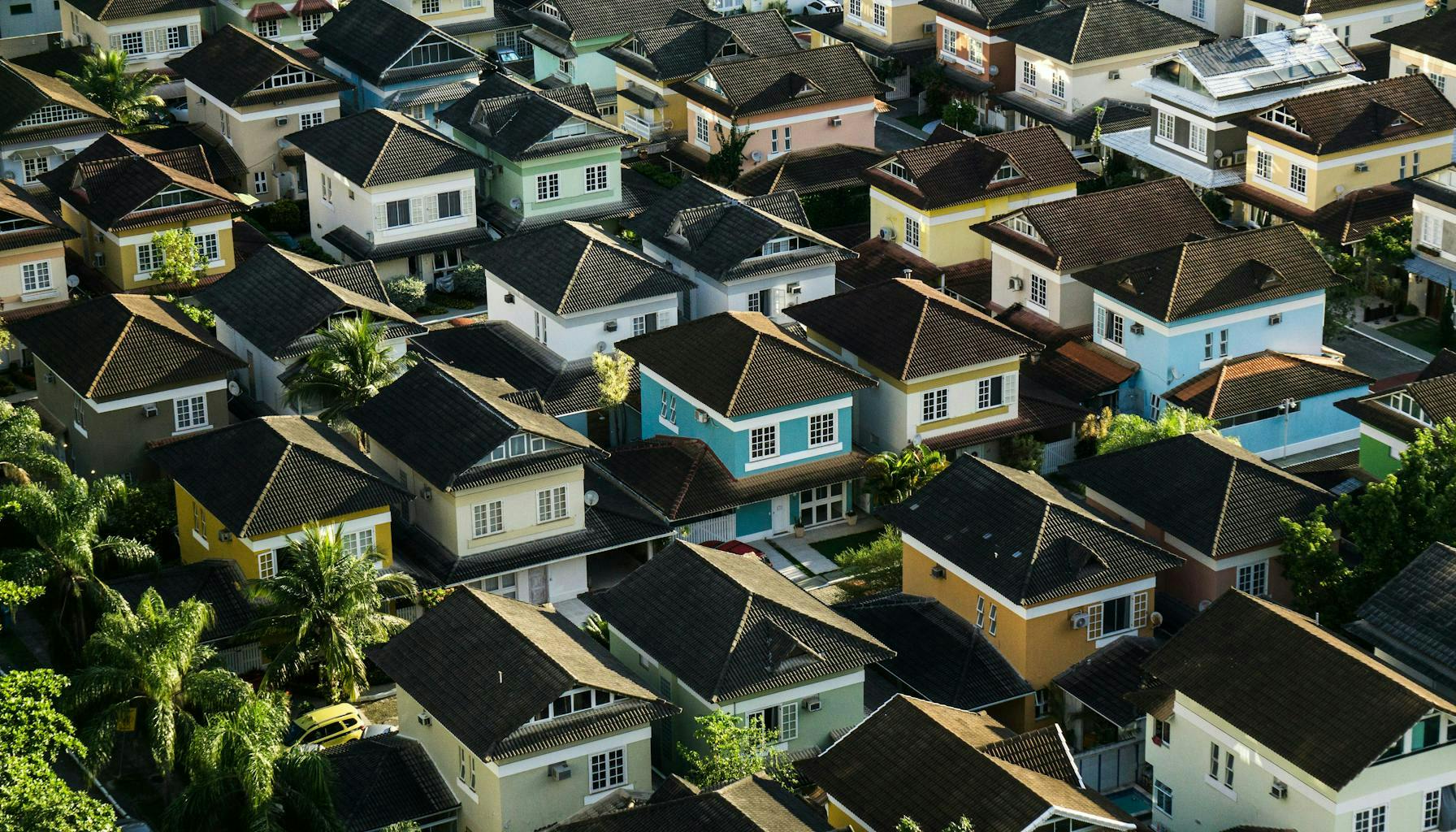 Photo by Breno Assis on Unsplash. Aerial view of a suburban neighborhood with neatly arranged, two-story houses painted in a variety of light colors like yellow, blue, and green. The homes have dark tiled roofs, and there are palm trees scattered throughout the area, contributing to a lush, tropical feel.