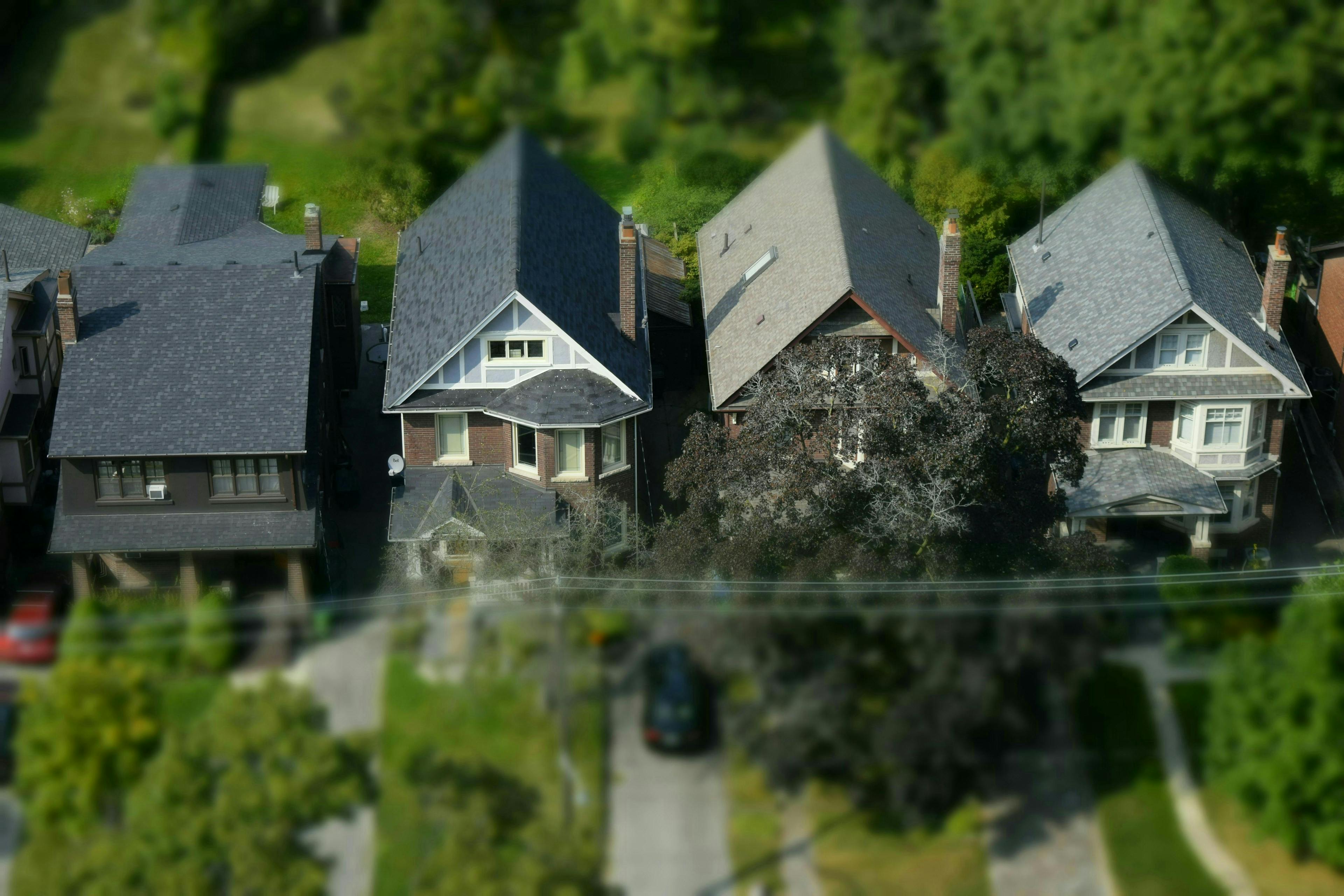 an aerial view of four houses lined up side by side in a residential neighborhood. The houses have steep, triangular roofs and are closely packed together with small yards in front. The houses vary slightly in design and roof color, with some having dark-colored roofs and others lighter. The surrounding area is green, with trees and lawns visible in the background, giving the scene a suburban feel.