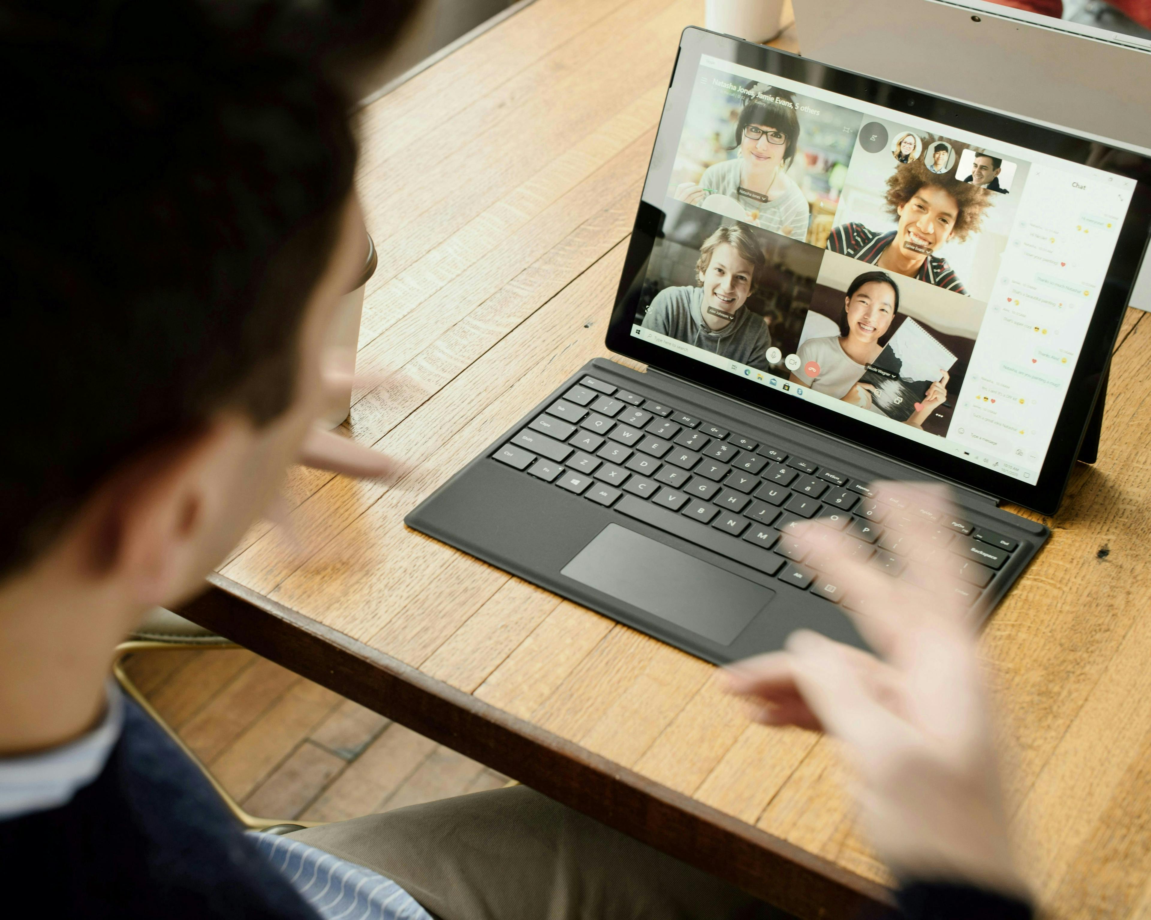 The image shows a person sitting at a wooden table, engaged in a video call on a tablet with a keyboard attached. The screen of the tablet displays a video conference with four participants, each in their own frame.