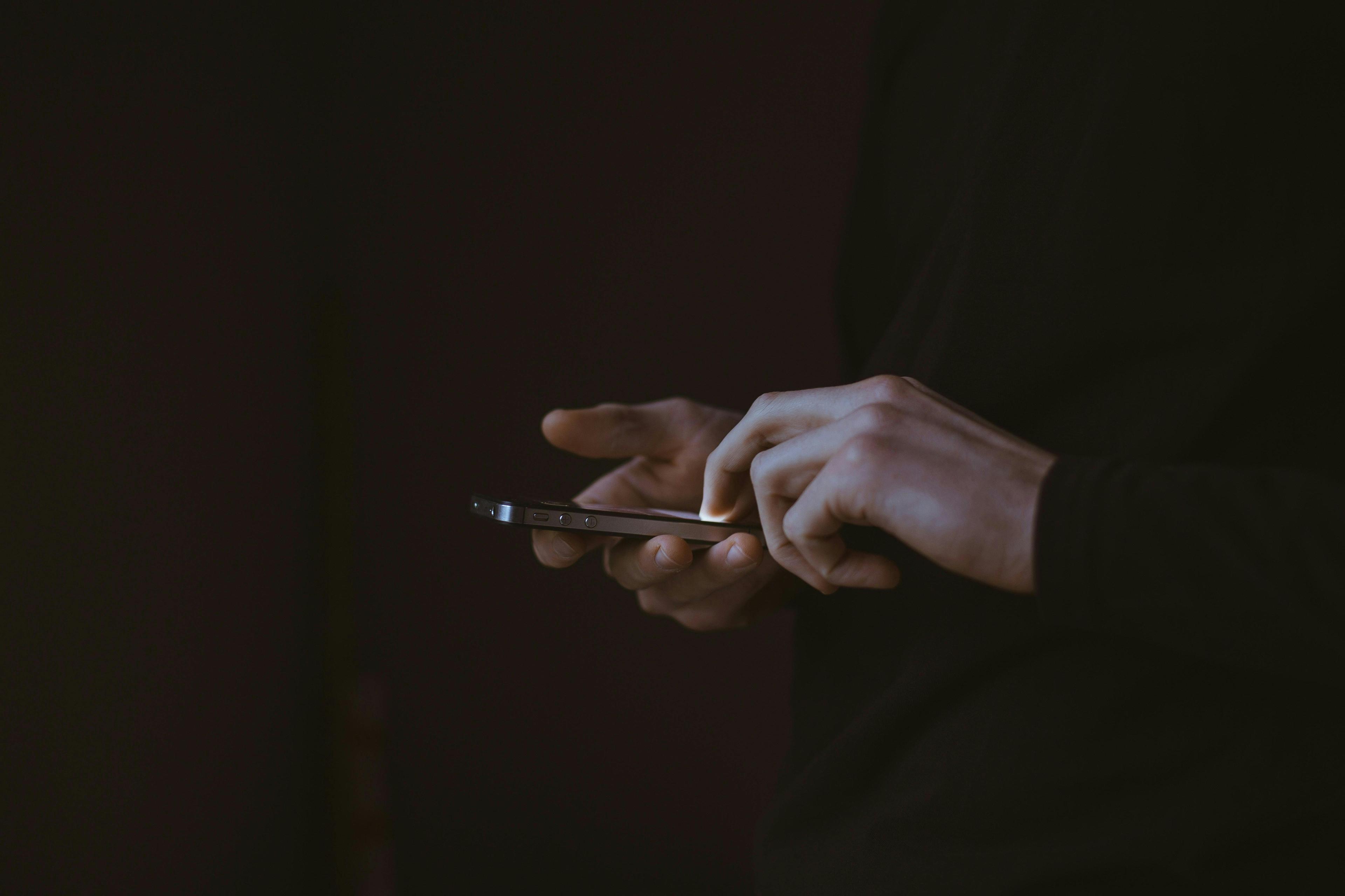 Close-up shot of a person using a smartphone. The individual is dressed in dark clothing, and only their hands and part of their phone are visible, set against a dark background.