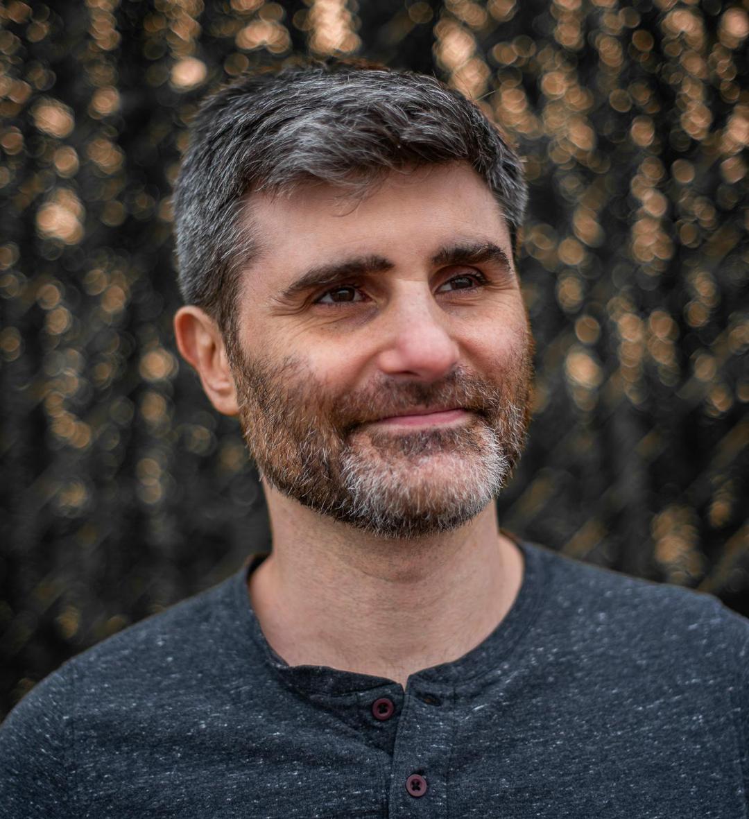 A man with short, neatly styled hair that is a mix of gray and black, complemented by a beard with similar tones. He is subtly smiling, wearing a dark gray shirt. The background features a soft bokeh effect with glimmers of light, giving the image a calm and warm atmosphere.