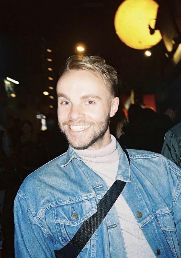 A smiling man with light hair and a beard is wearing a denim jacket over a light turtleneck. He is standing in a nighttime setting, with warm lights glowing in the background, including a large, glowing yellow sphere. He has a black strap across his chest, possibly from a bag, and the environment around him suggests an outdoor, urban atmosphere.