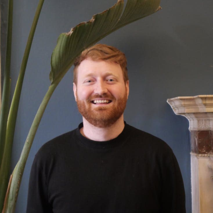 A person with short red hair and a beard is smiling in front of a dark blue wall. There is a large green plant and a decorative architectural element in the background.