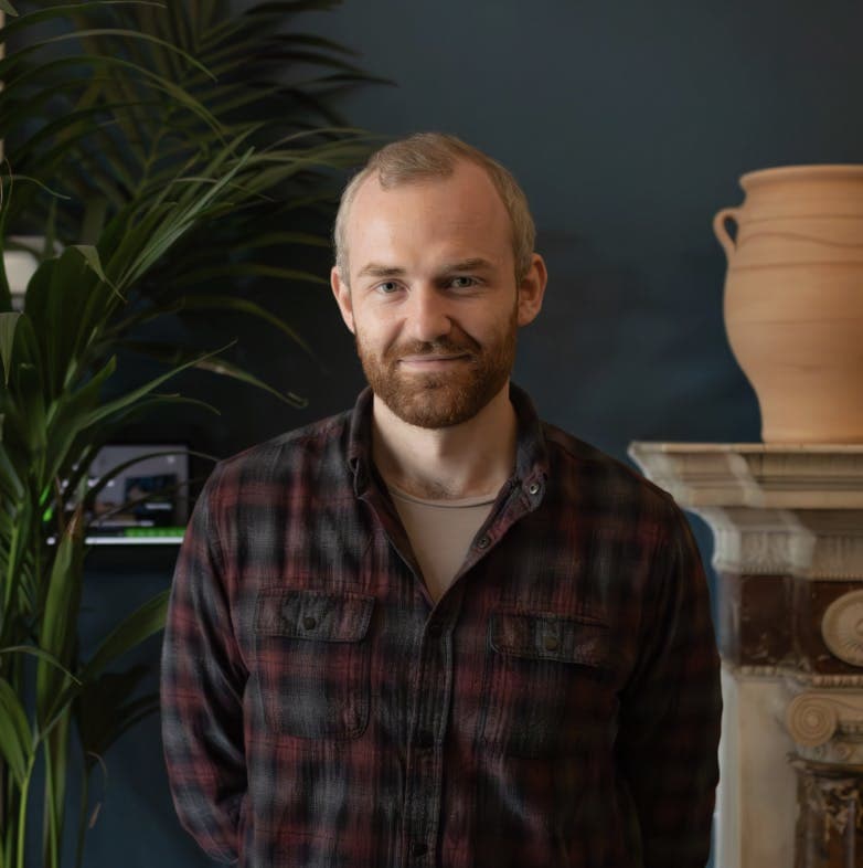 A person with short light brown hair and a beard is standing in front of a dark blue wall, wearing a plaid shirt. There is a green plant and a decorative ceramic vase on a mantelpiece in the background, creating a warm and cozy atmosphere.