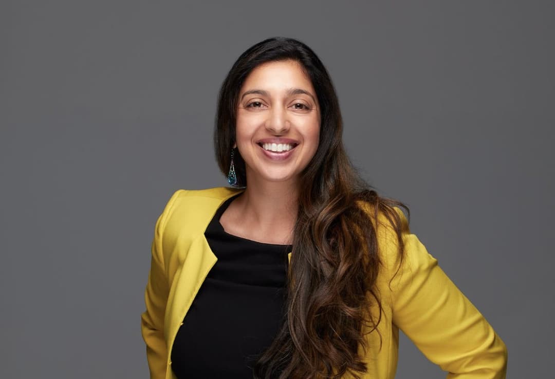 The image shows a woman with long dark hair wearing a bright yellow jacket over a black top, smiling confidently. She is posing against a plain gray background, and her expression is warm and approachable.