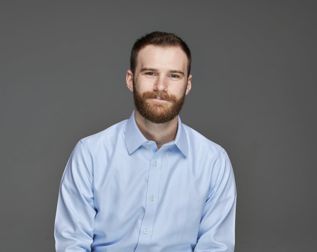 A man with a beard, wearing a light blue button-up shirt, posing against a plain gray background. He has a calm and friendly expression.