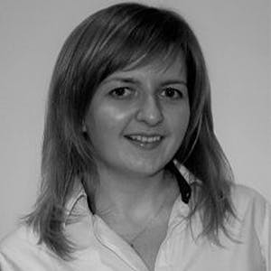 A woman, slightly smiling, is facing the camera while wearing a light-colored shirt, with straight hair and a neutral background.