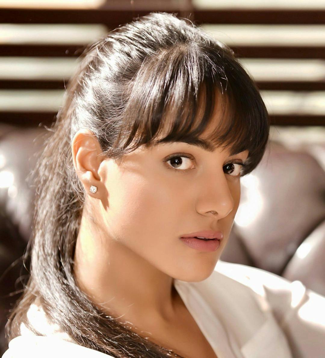 A woman with straight, dark hair and bangs, wearing a white top and earrings, looks directly at the camera in a brightly lit room with brown furniture and slatted blinds.