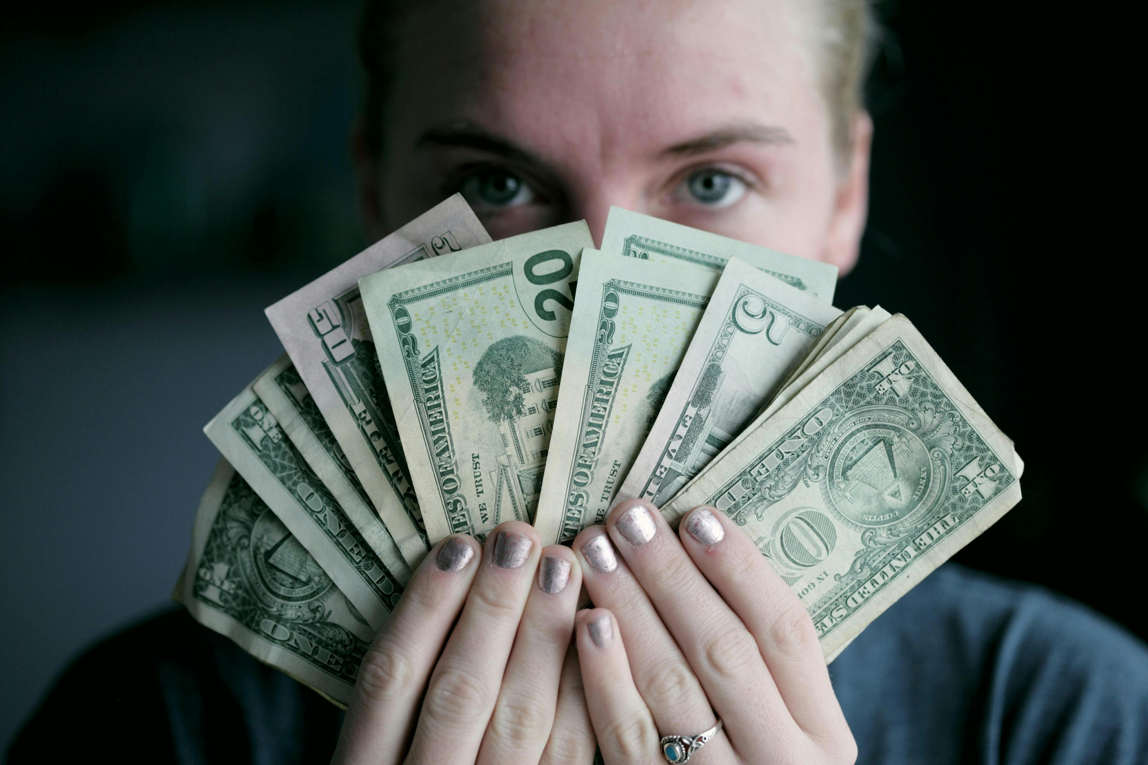 Hands fanning out several US dollar bills in multiple denominations, partially covering a person’s face in the background. The context is dark and indistinct.