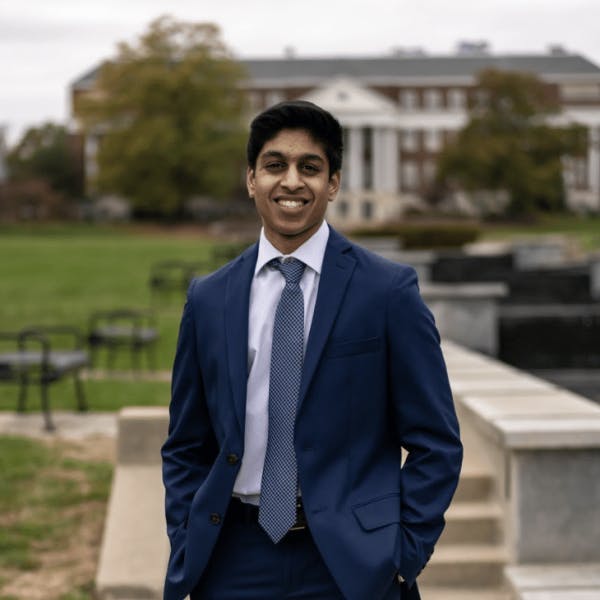 A person in a blue suit, white shirt, and blue tie stands smiling with hands in pockets on a pathway with a grassy area and a large, white-columned building in the background.