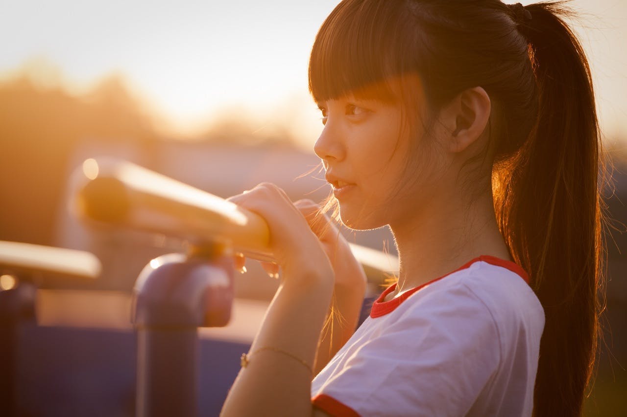 A young woman with a ponytail leans on a horizontal bar outdoors, gazing into the distance during a golden-hued sunset. She wears a white T-shirt with red trim.