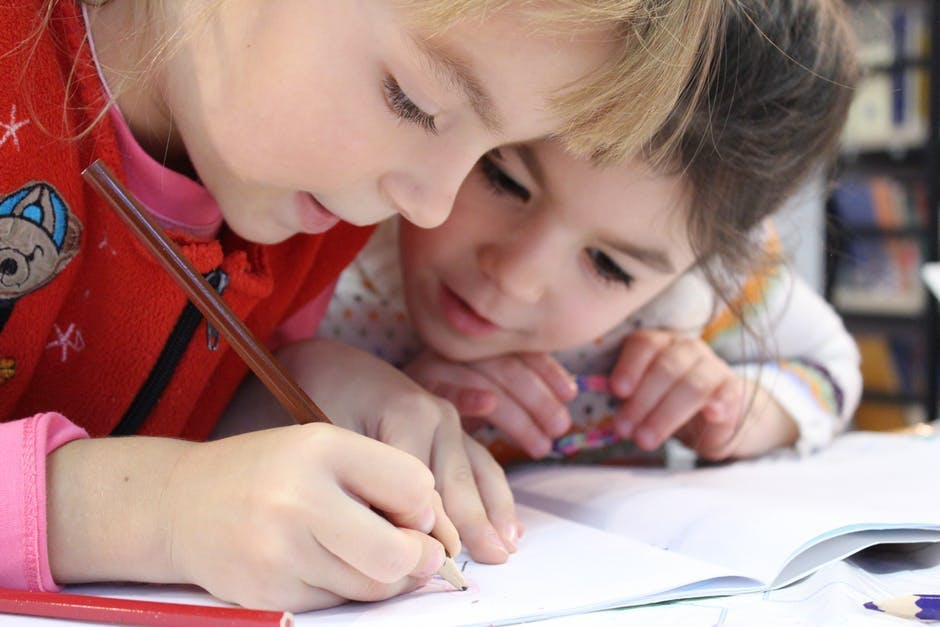 Two children drawing with colored pencils on paper, their heads close together; one child focused on drawing, while the other watches intently, in a bright indoor setting.