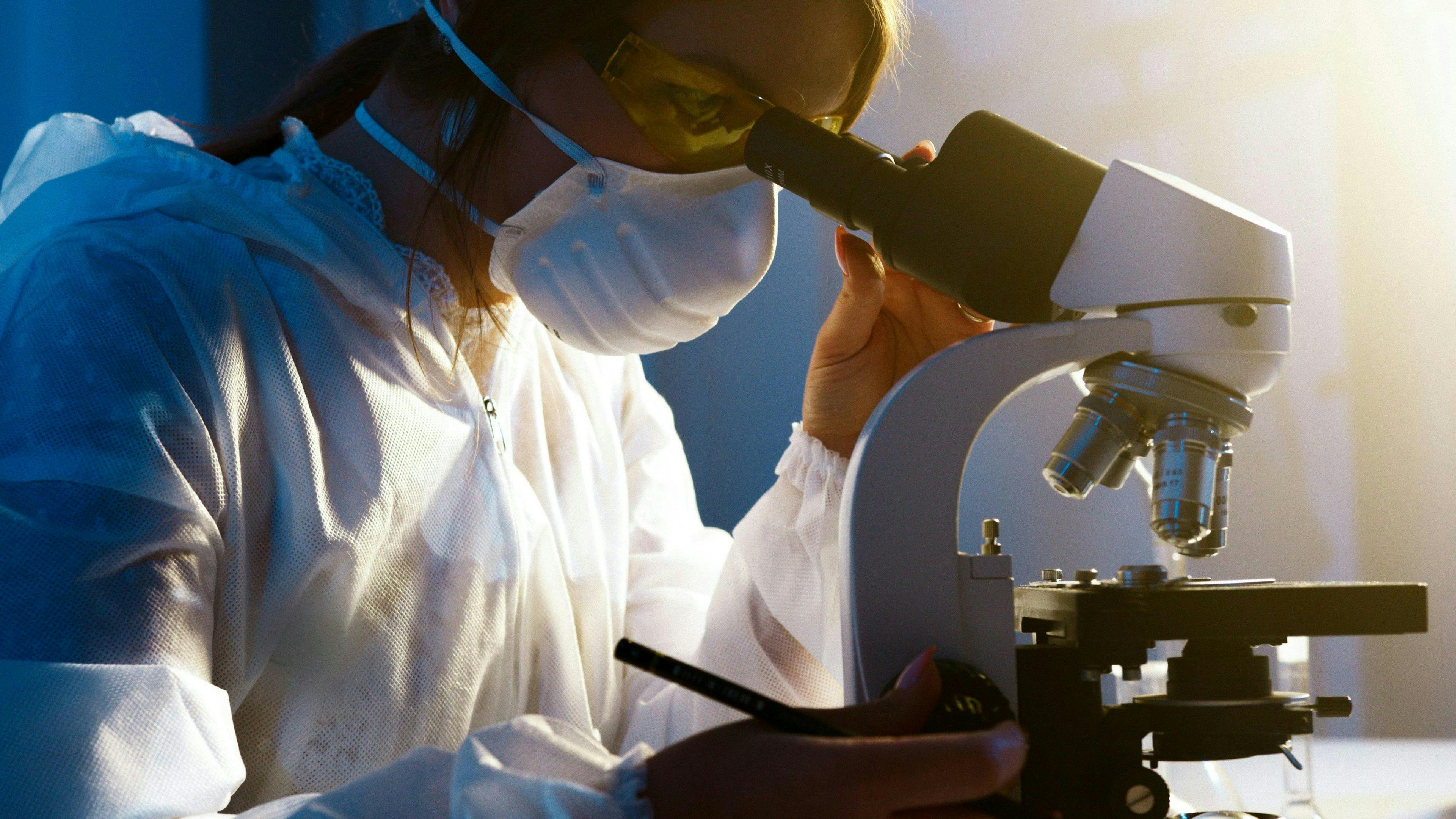 A scientist wearing protective gear and a mask examines a sample through a microscope in a brightly lit laboratory.