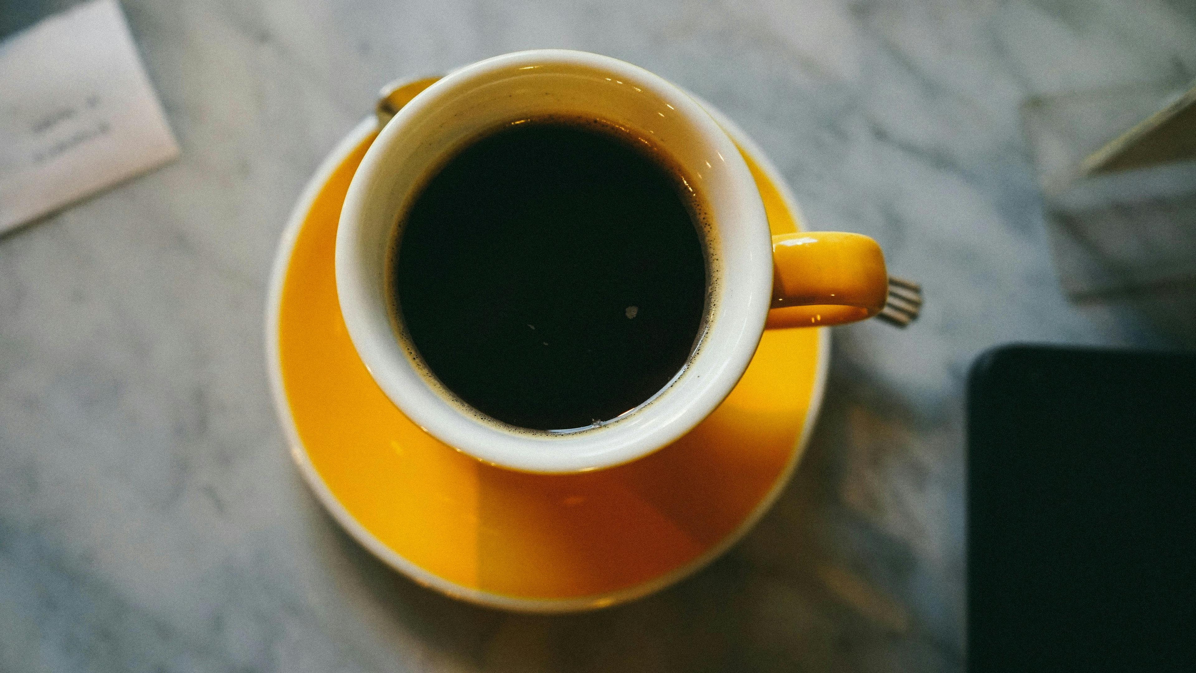 A yellow cup filled with black coffee sits on a matching saucer on a marble surface, with part of a piece of paper and device visible nearby.