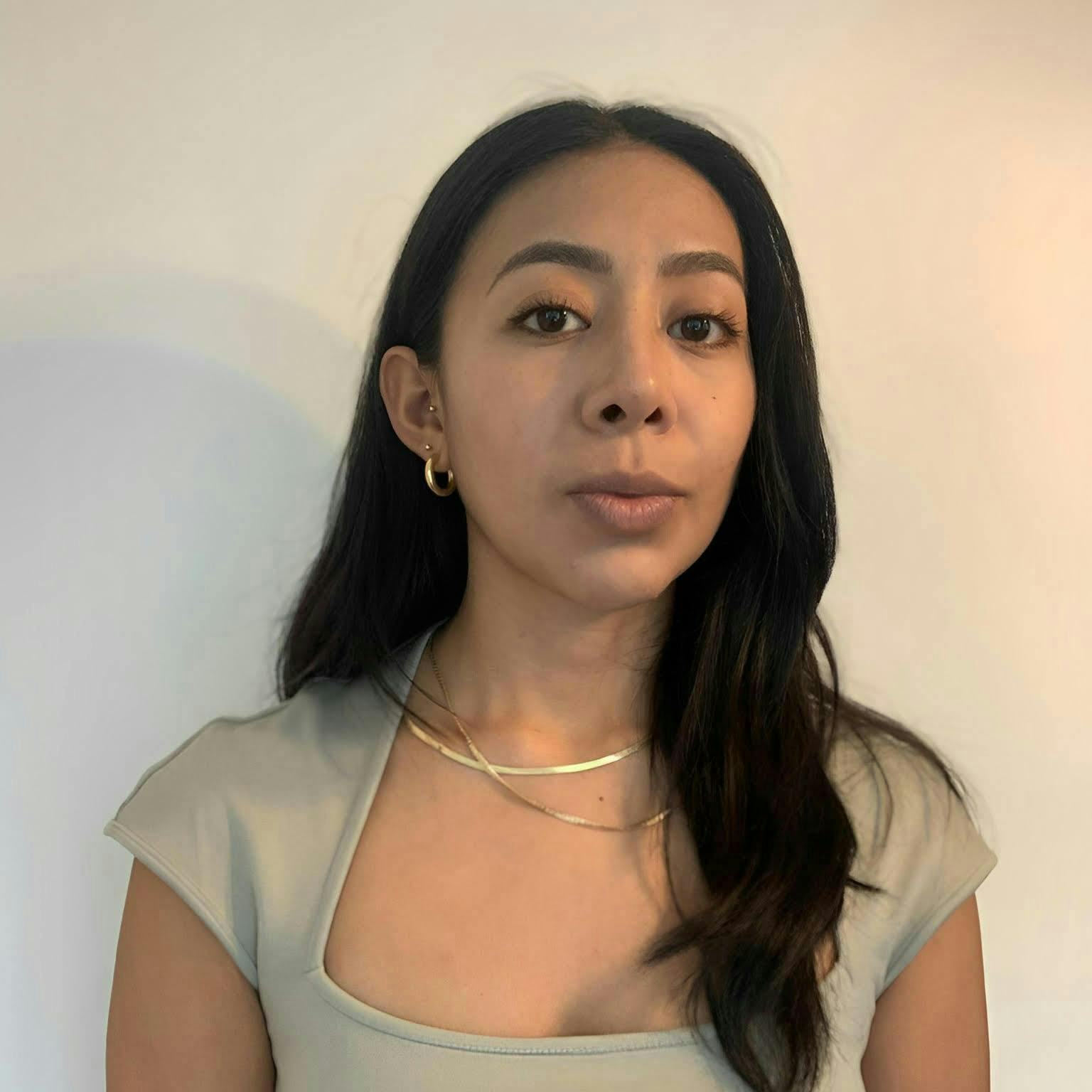 A woman with long black hair wears a light gray top, gold hoop earrings, and layered gold necklaces, standing against a plain, light-colored background, gazing directly at the camera.