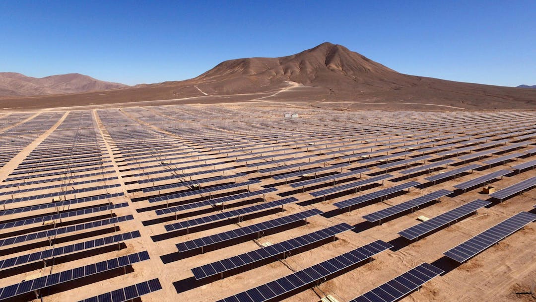 Solar panels are arranged in rows, harnessing sunlight in a vast, arid desert landscape with large, barren mountains in the background under a clear blue sky.
