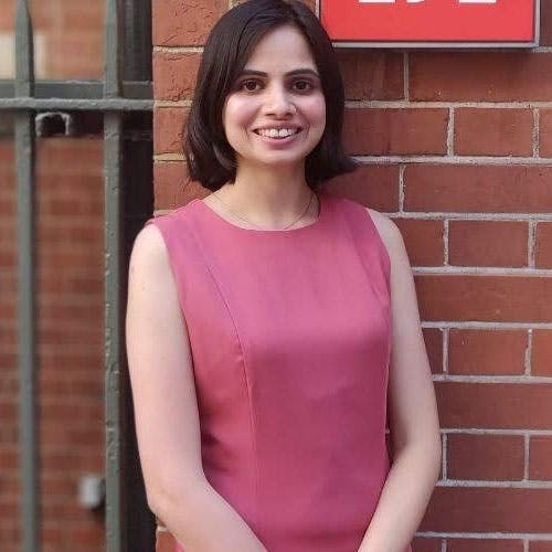 A smiling woman in a sleeveless pink dress stands against a red brick wall next to a metal fence. Part of a red sign is visible in the top right corner.