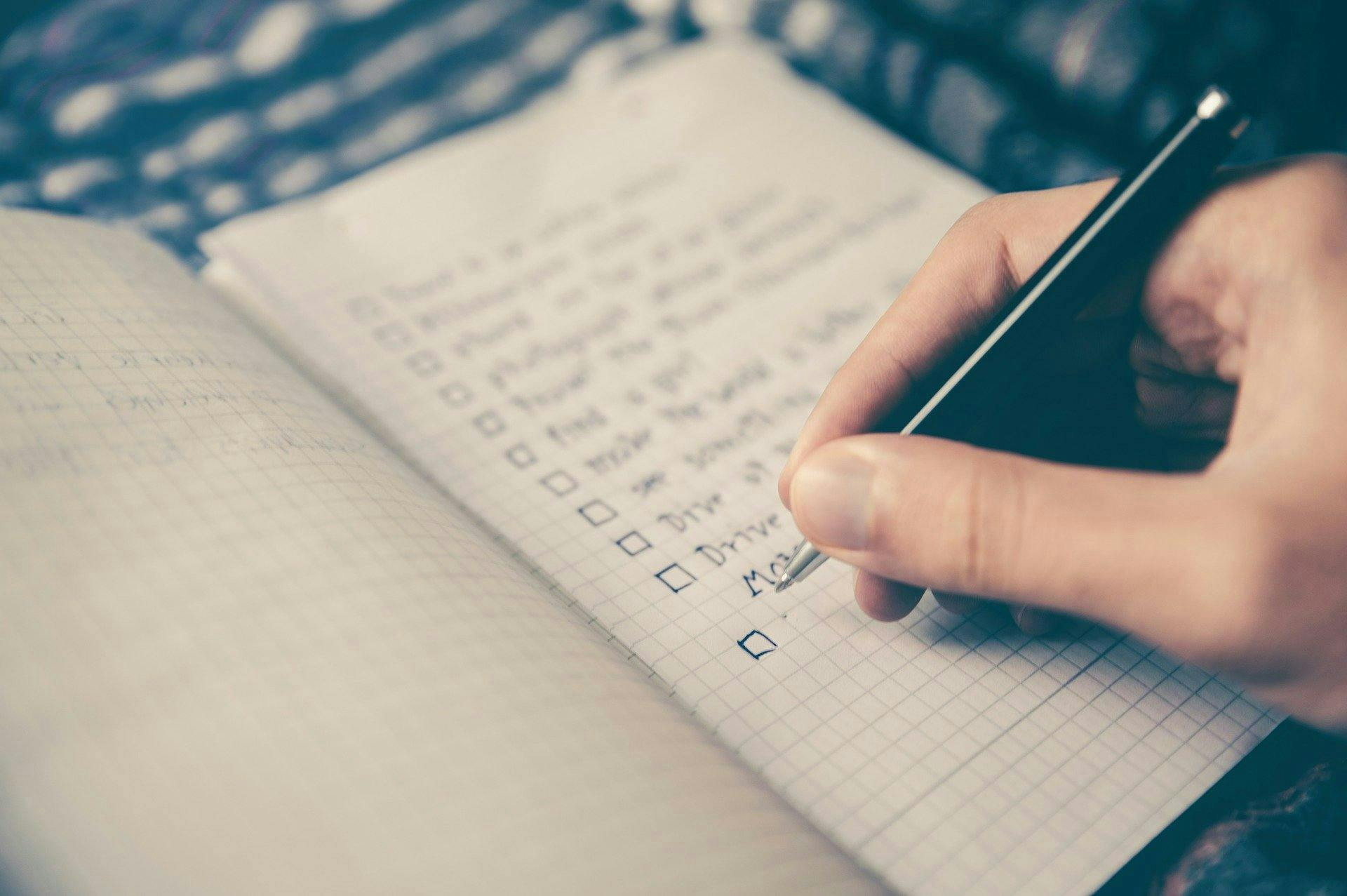 A hand writes a checklist in a grid-style notebook, beside an open second notebook on a patterned surface.