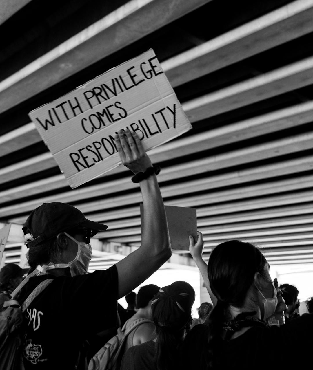 A person holds a sign reading "WITH PRIVILEGE COMES RESPONSIBILITY" during a protest under a bridge, surrounded by a crowd of people wearing masks and caps.