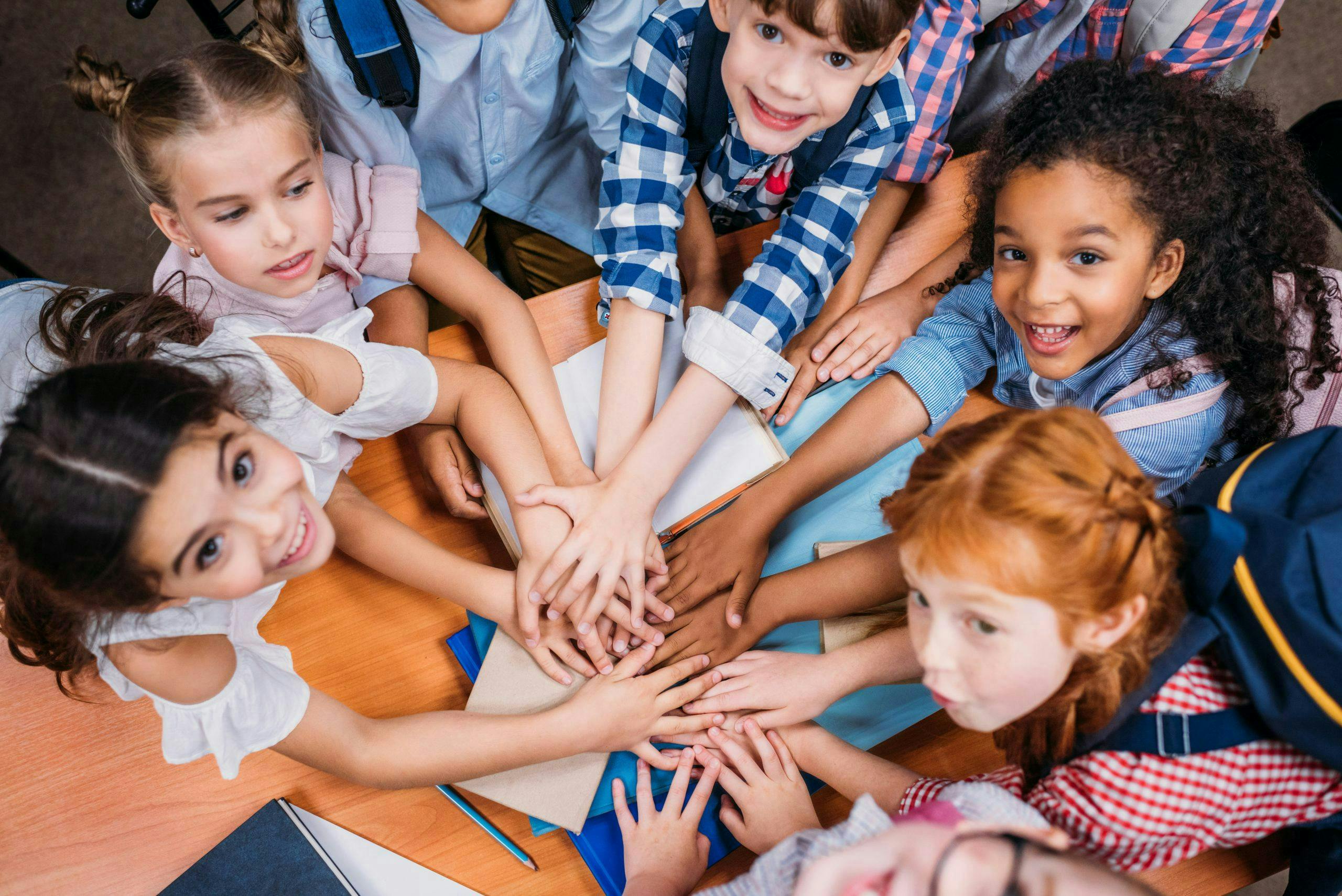 Children place their hands together in a group gesture atop a wooden desk crowded with books and notebooks, displaying unity and teamwork in a classroom setting.