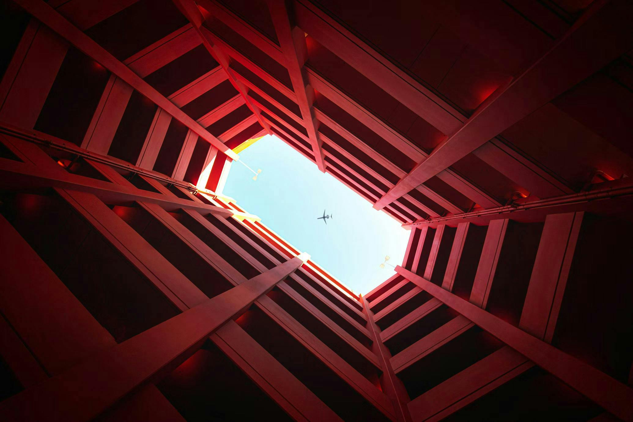 An airplane flies overhead, visible through a symmetrical red architectural structure's opening, with sunlight illuminating the edges, set against a clear blue sky.