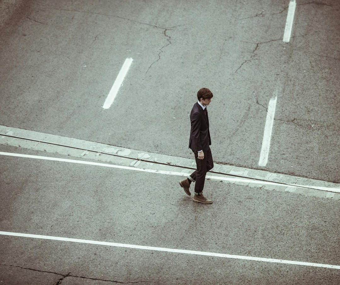 A person in a suit and brown shoes walks across a deserted, cracked street marked with faded white lines.